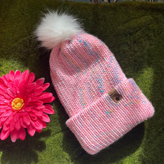 A pink multi-colored beanie with a white faux fur pom-pom on a green background, with a custom cork tag attached to the brim. spring chunky beanie in pink