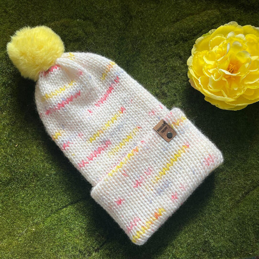A knitted beanie in creamy white with yellow, dark pink, and periwinkle specks, featuring a faux fur pom-pom and a custom cork tag on a green background with a yellow flower. basic beanie in spring sprinkle