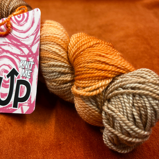 A skein of KnitMeUp 100% Merino Wool yarn in the hand-painted color "Tim," featuring warm orange and brown tones. Displayed partially unwound on a neutral background. close up