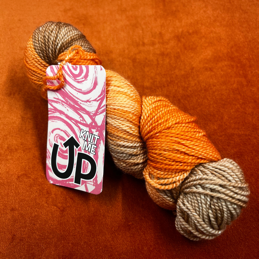 A skein of KnitMeUp 100% Merino Wool yarn in the hand-painted color "Tim," featuring warm orange and brown tones. Displayed partially unwound on a neutral background.