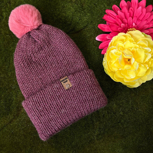 A dark pink, double-layered wooly beanie with a faux fur pom-pom on a green background, next to a yellow flower. rose seduction beanie dark pink the wooly beanie