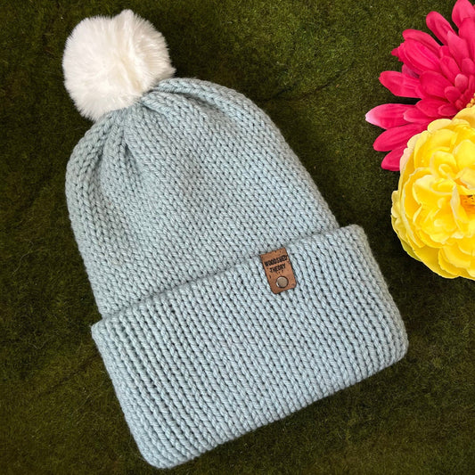 A knitted beanie in ice blue with a white faux fur pom-pom on a green background, next to a pink and yellow flower. the basic beanie in ice blue flat front