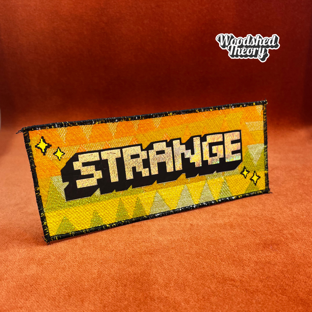 “That’s Strange iron-on patch with holographic pixel font and vintage orange, yellow, and green triangle background – handmade by Woodshed Theory.” front