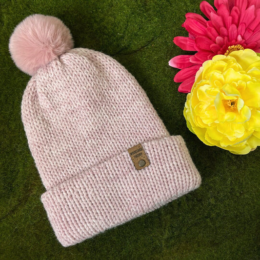 A pale pink wooly beanie with a pom-pom on a green background, next to a yellow and a pink artificial flower. the wooly beanie in pale pink with pink pom pom front flat