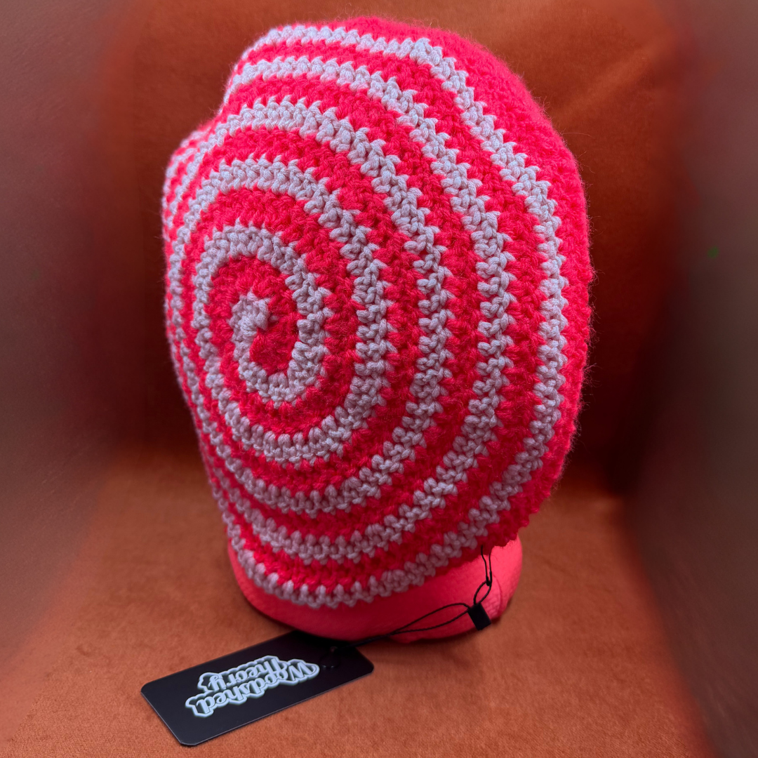 A crocheted spiral hat in bright pink and light gray, displayed on a brown textured surface with a black 'Local Treasure' tag in the foreground.