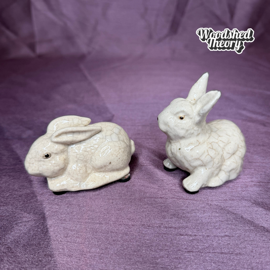 Two small ceramic rabbit figurines in an off-white glaze, displayed on a textured purple fabric background. A 'Woodshed Theory' brand logo is visible in the top right corner.