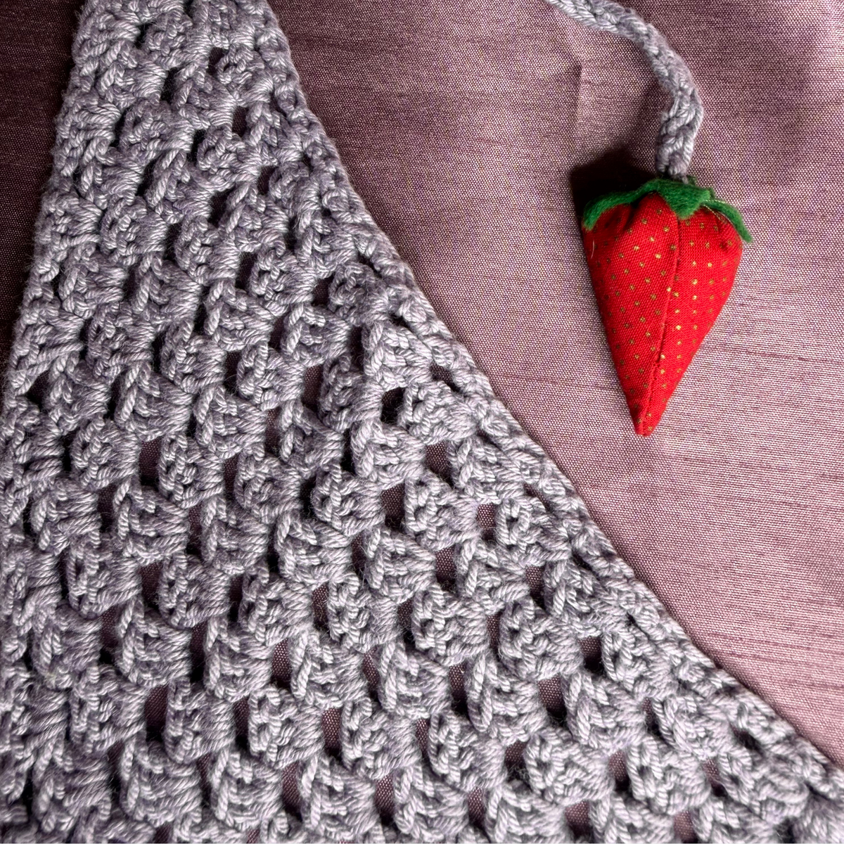 A close-up of a gray crocheted textile with an openwork knit pattern, laid on a mauve fabric background and accented by a small red strawberry-shaped charm with green leaves.