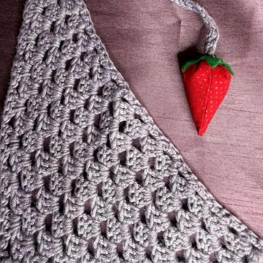 A close-up of a gray crocheted textile with an openwork knit pattern, laid on a mauve fabric background and accented by a small red strawberry-shaped charm with green leaves.
