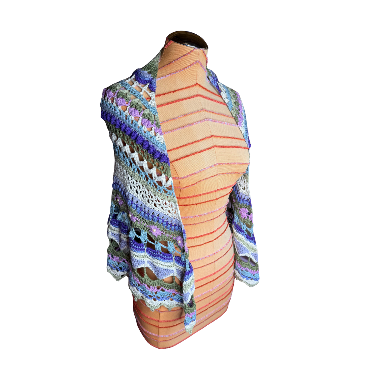A multicolor crochet cardigan with blue, purple, green, and orange patterns, draped on a peach-colored dress form mannequin against a white background.