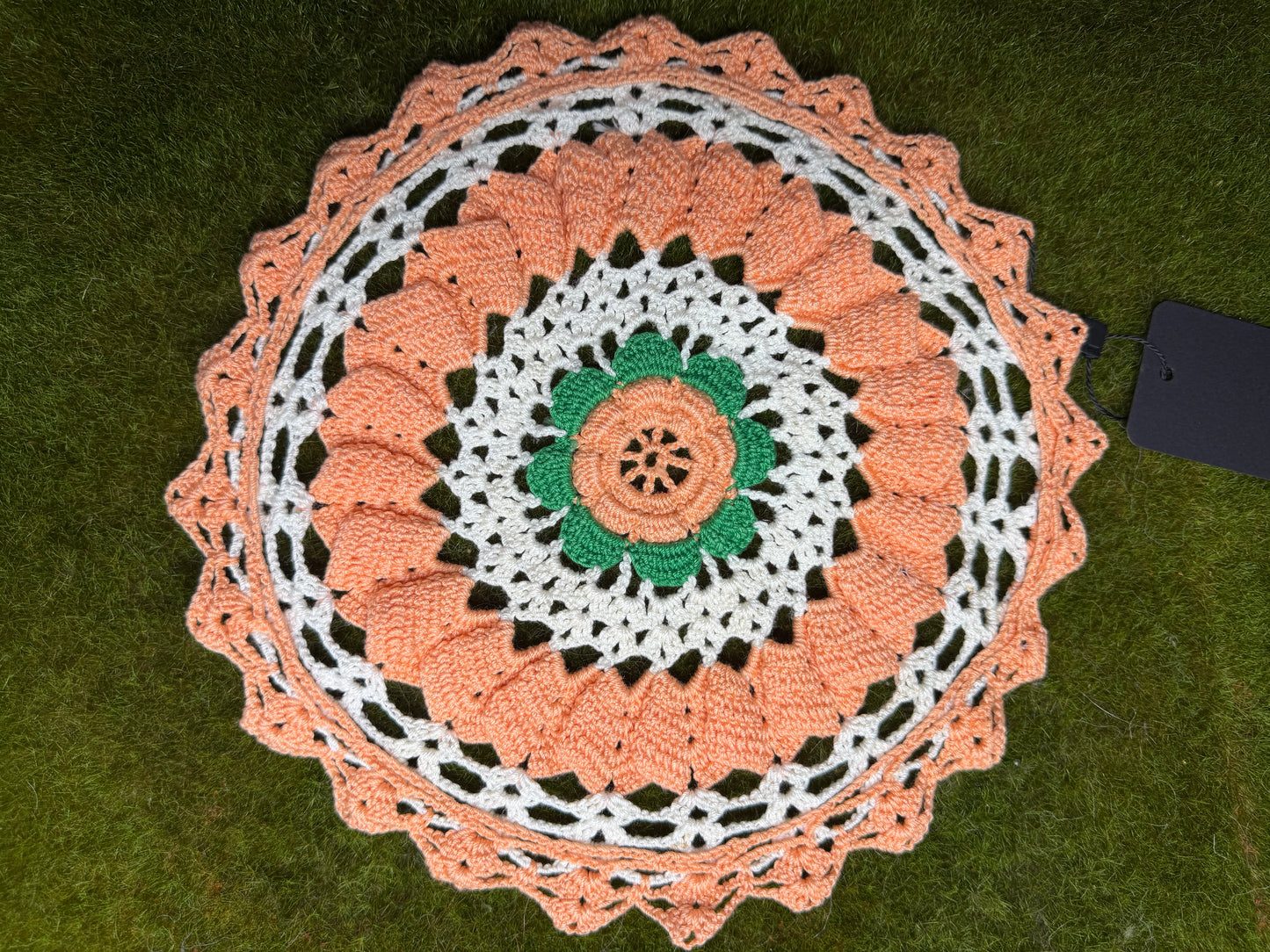 A crocheted round doily with a scalloped edge, featuring concentric rings of peach, white, and green crochet patterns, laid flat on green grass next to a black tag. back