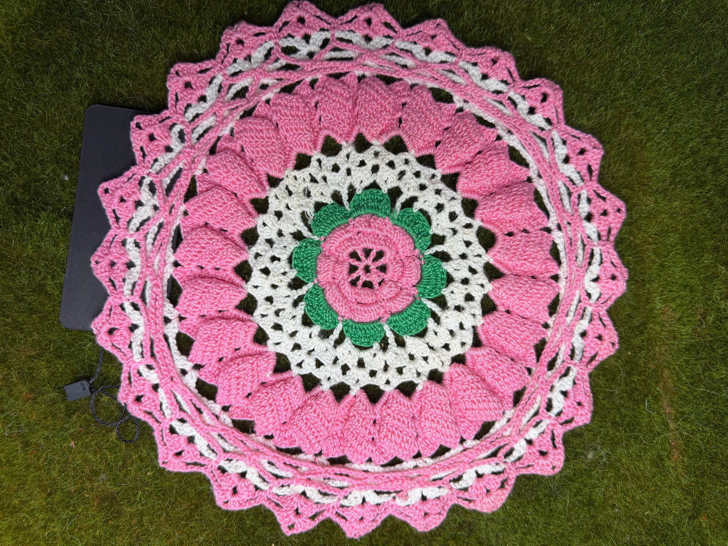 A round crocheted doily with a pink and white floral mandala pattern and a green center, laid flat on green grass next to a black laptop.