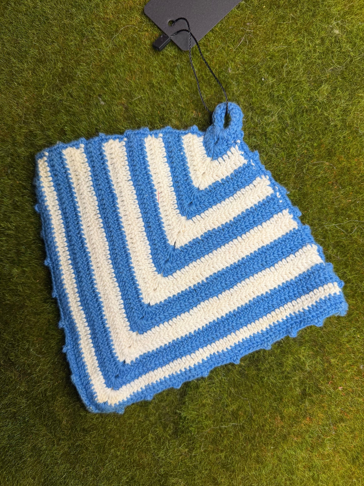 A crocheted square textile item with alternating blue and off-white stripes, laid flat on green grass. A small black tag is attached at one corner.