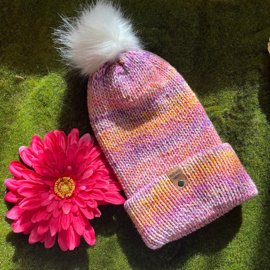 A multicolored knitted beanie with a faux fur pom-pom on a green surface, next to a pink flower. fairy party front