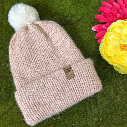 A nude pink alpaca and acrylic beanie with a pom-pom on a green background, with a custom faux cork tag attached. Alpaca Snow Beanie Alpaca Snow Beanie Nude Pink
