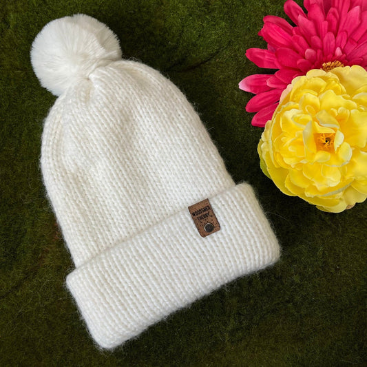 A white alpaca and acrylic knit beanie with a pom-pom, displayed on a green background with a yellow and pink flower to the side. alpaca snow beanie in snow hare bright weight with logo tag and removable pom pom front