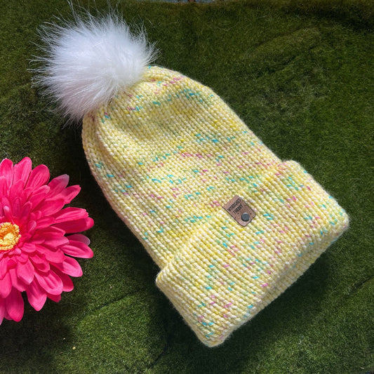 A yellow and multicolored beanie with a faux fur pom-pom on a green background, with a custom cork tag attached to it. woodshed theory spring beanie yellow