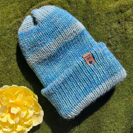A striped blue and gray beanie with a custom cork tag displaying the Woodshed Theory logo, placed on a grassy surface beside a yellow flower. Muted blue double-layered knit beanie with cork tag, made with organic cotton, wool, and recycled fiber, hand crafted by Woodshed Theory