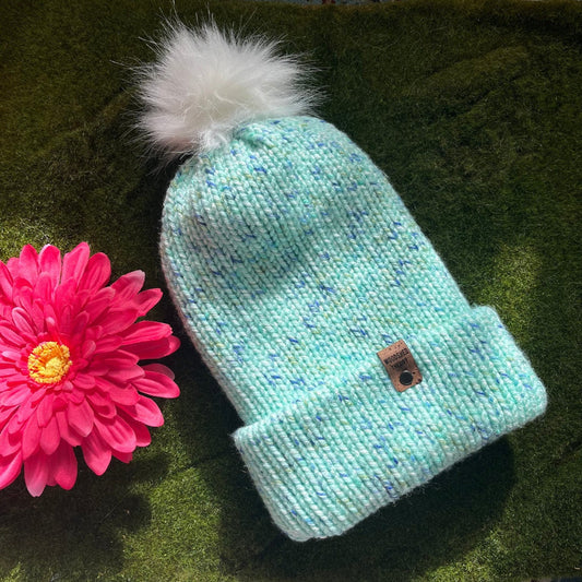 A teal blue chunky knit beanie with a faux fur pom-pom on a green background, next to a pink flower. front