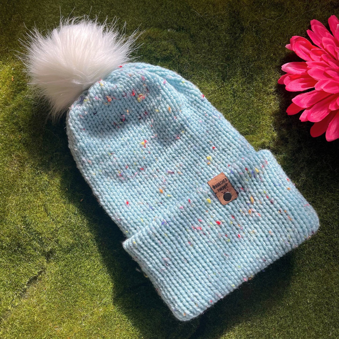 A light blue beanie with multicolor specks and a faux fur pom-pom on a green background, with a custom cork tag attached to the brim. basic beanie in robins egg flat front