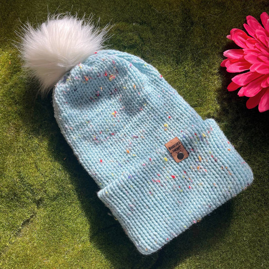 A light blue beanie with multicolor specks and a faux fur pom-pom on a green background, with a custom cork tag attached to the brim. basic beanie in robins egg flat front
