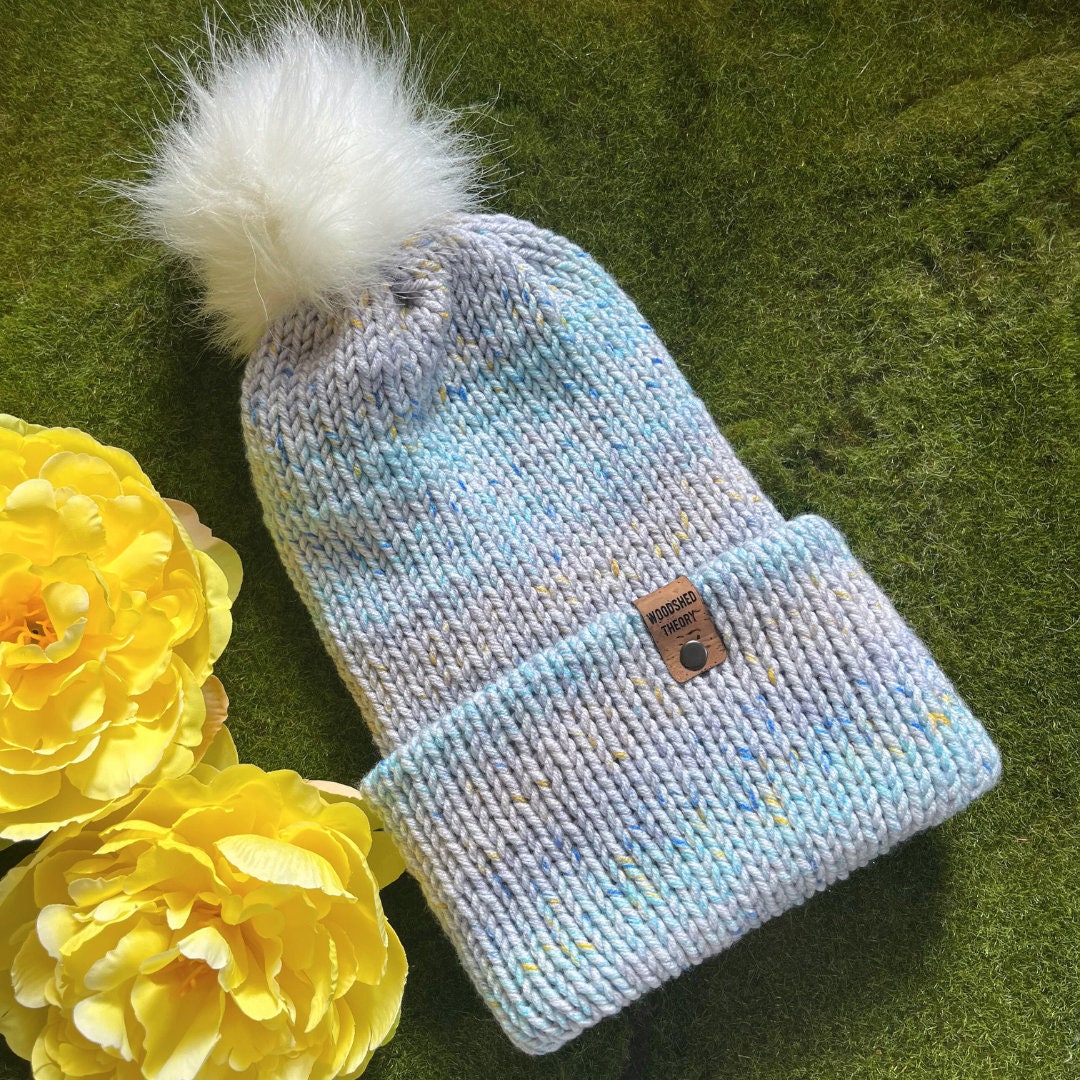 A blue and white speckled beanie with a white faux fur pom-pom, displayed on a green surface with yellow flowers to the side. The beanie features a custom cork tag with the Woodshed Theory logo.