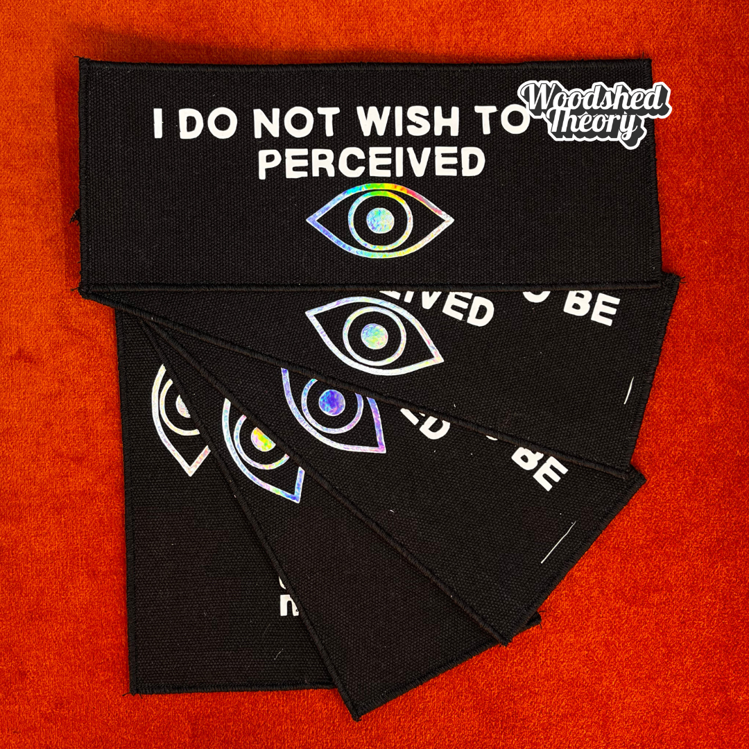 Black iron-on canvas patch with glow-in-the-dark white text reading “I do not wish to be perceived” and a holographic eye illustration. Approx. 3x8 inches.