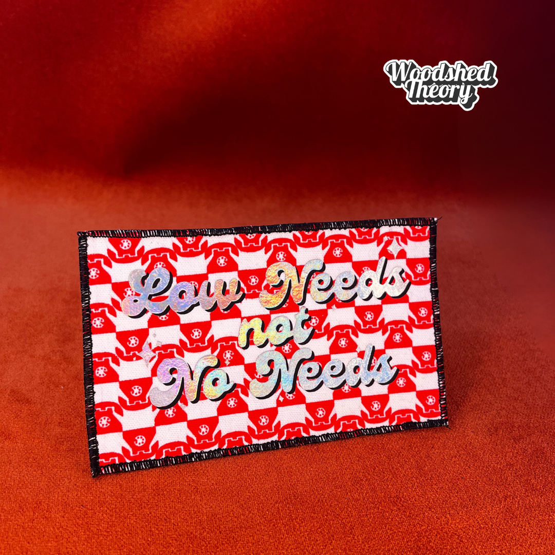 Rectangular iron-on patch reading "Low Needs, Not No Needs" in clean bold type. Made with fabric and backing, approx. 5.25 x 3 inches. front halographic red telephones 


