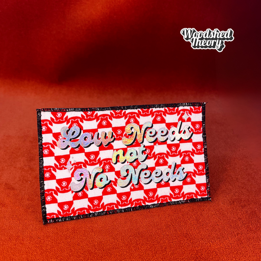 Rectangular iron-on patch reading "Low Needs, Not No Needs" in clean bold type. Made with fabric and backing, approx. 5.25 x 3 inches. front halographic red telephones 


