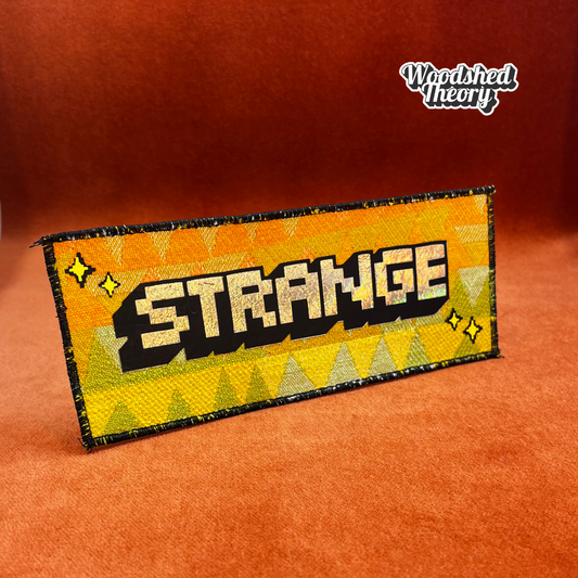 “That’s Strange iron-on patch with holographic pixel font and vintage orange, yellow, and green triangle background – handmade by Woodshed Theory.” front