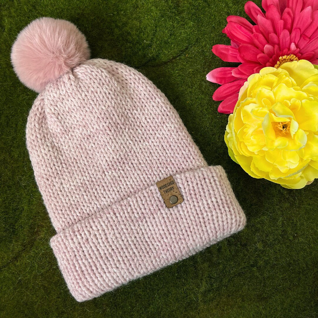 A pale pink wooly beanie with a pom-pom on a green background, next to a yellow and a pink artificial flower. the wooly beanie in pale pink with pink pom pom front flat 