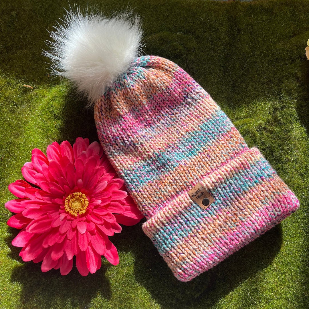 A multicolored knitted beanie with a faux fur pom-pom on a green surface, next to a pink flower. woodshed theory flat