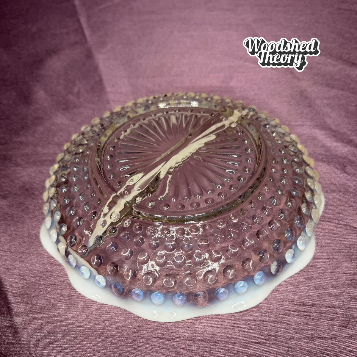 A vintage Fenton hobnail glass bowl in a dusty purple shade, featuring a white scalloped base and blue glass beads along the edge, displayed on a textured purple fabric background.