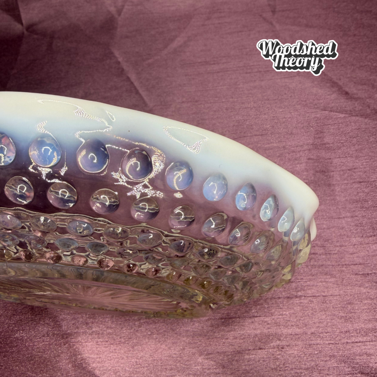 A close-up of a ribbed glass bowl with a milky white top and iridescent blue-purple tones, resting on a textured mauve fabric background. The Woodshed Theory logo is visible in the upper right.