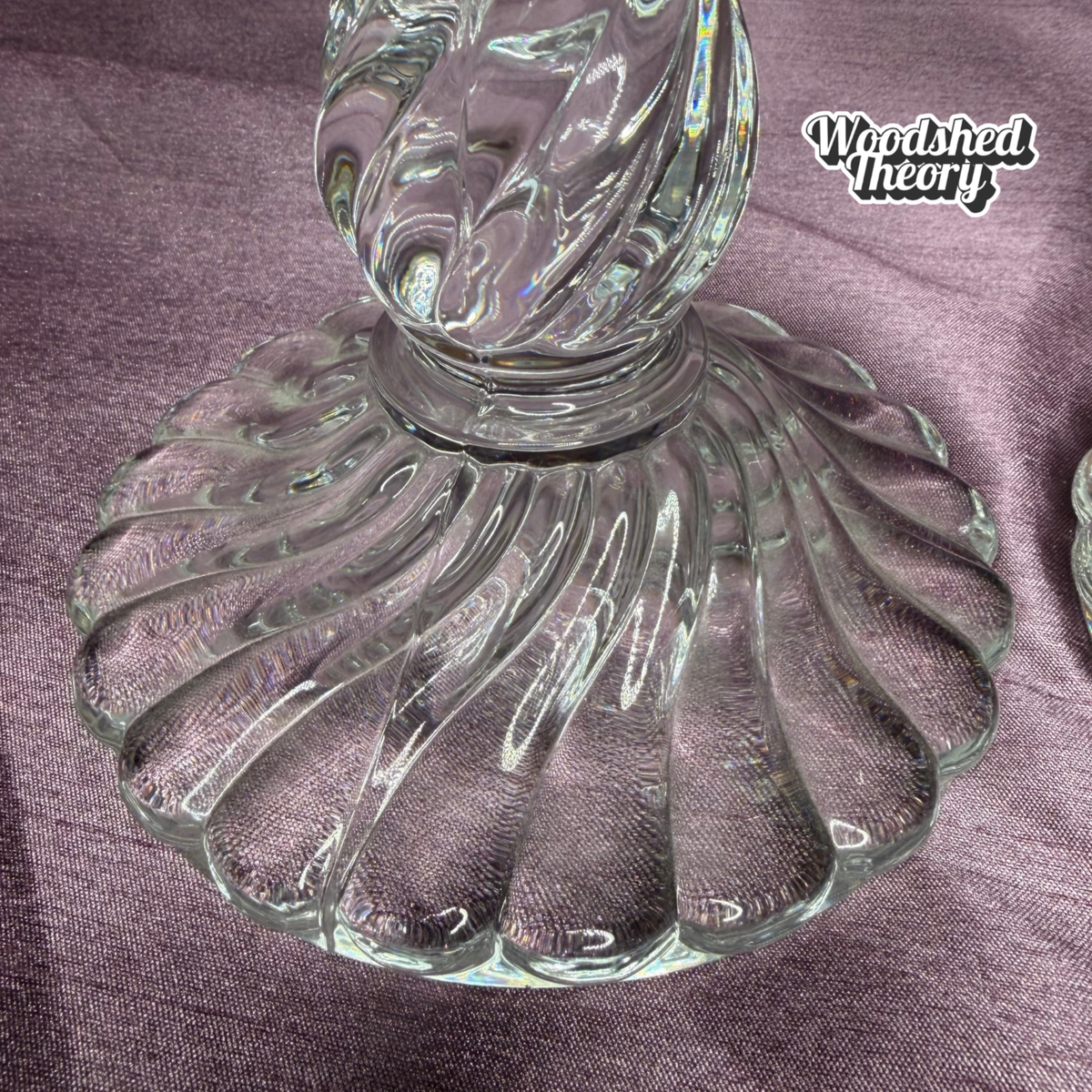 A close-up of a clear ribbed glass object, likely a candlestick base or decorative glass piece, resting on a textured purple fabric background. A 'Woodshed Theory' logo is visible in the top right corner.