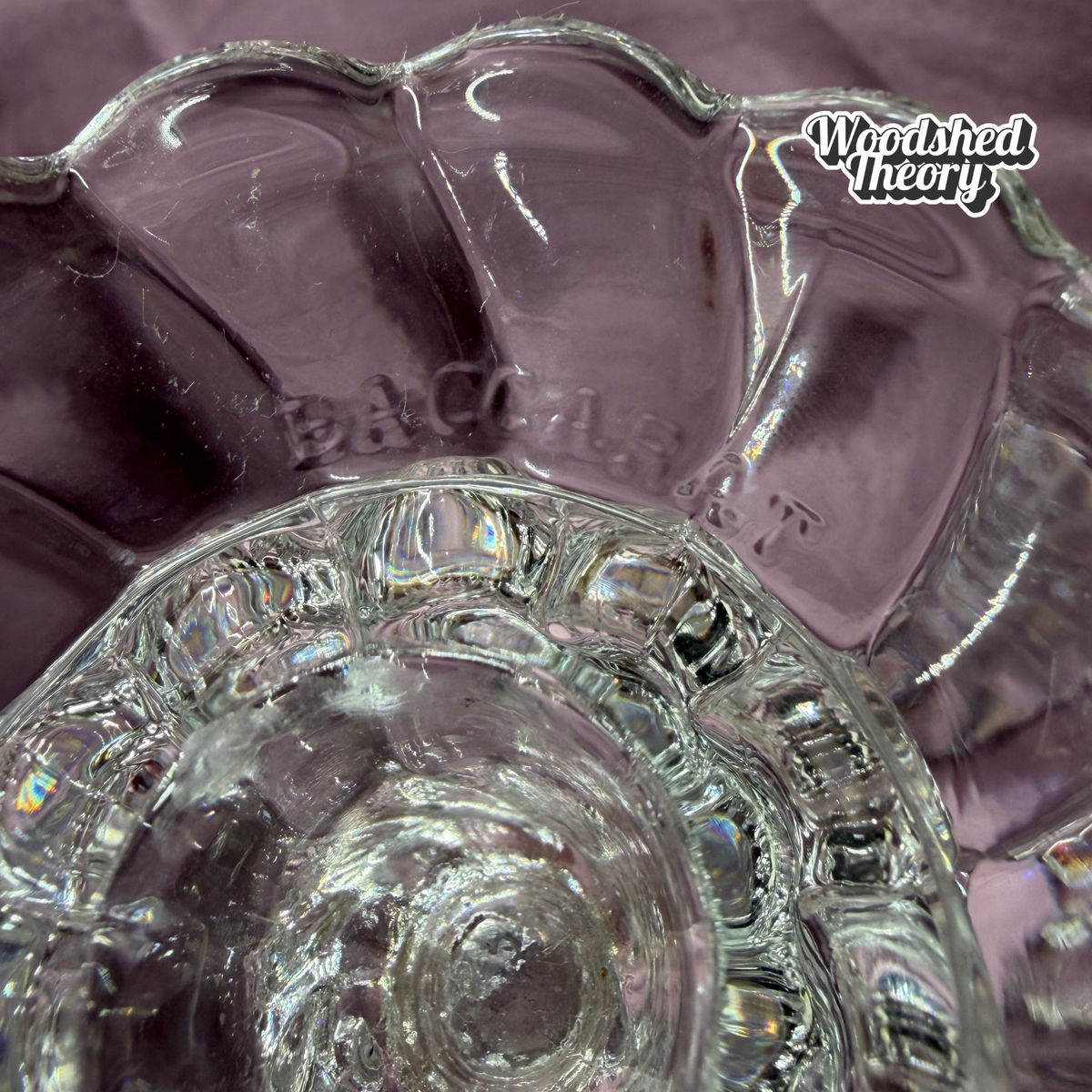 A close-up of a clear glass bowl or dish with a scalloped rim, showing embossed lettering and a textured ribbed base, photographed against a soft lavender-purple fabric background. A 'Woodshed Theory' watermark is visible in the top right.