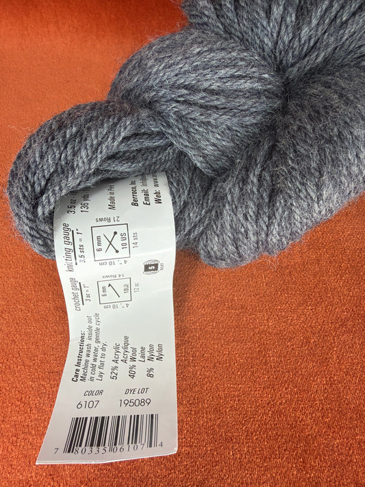 A close-up of a gray heathered yarn skein with a white product label attached, placed on a textured orange-brown surface.