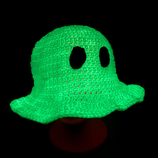 A bright neon green crocheted ghost hat with two large eye cutouts and a stitched mouth, displayed on a dark stand against a black background.
