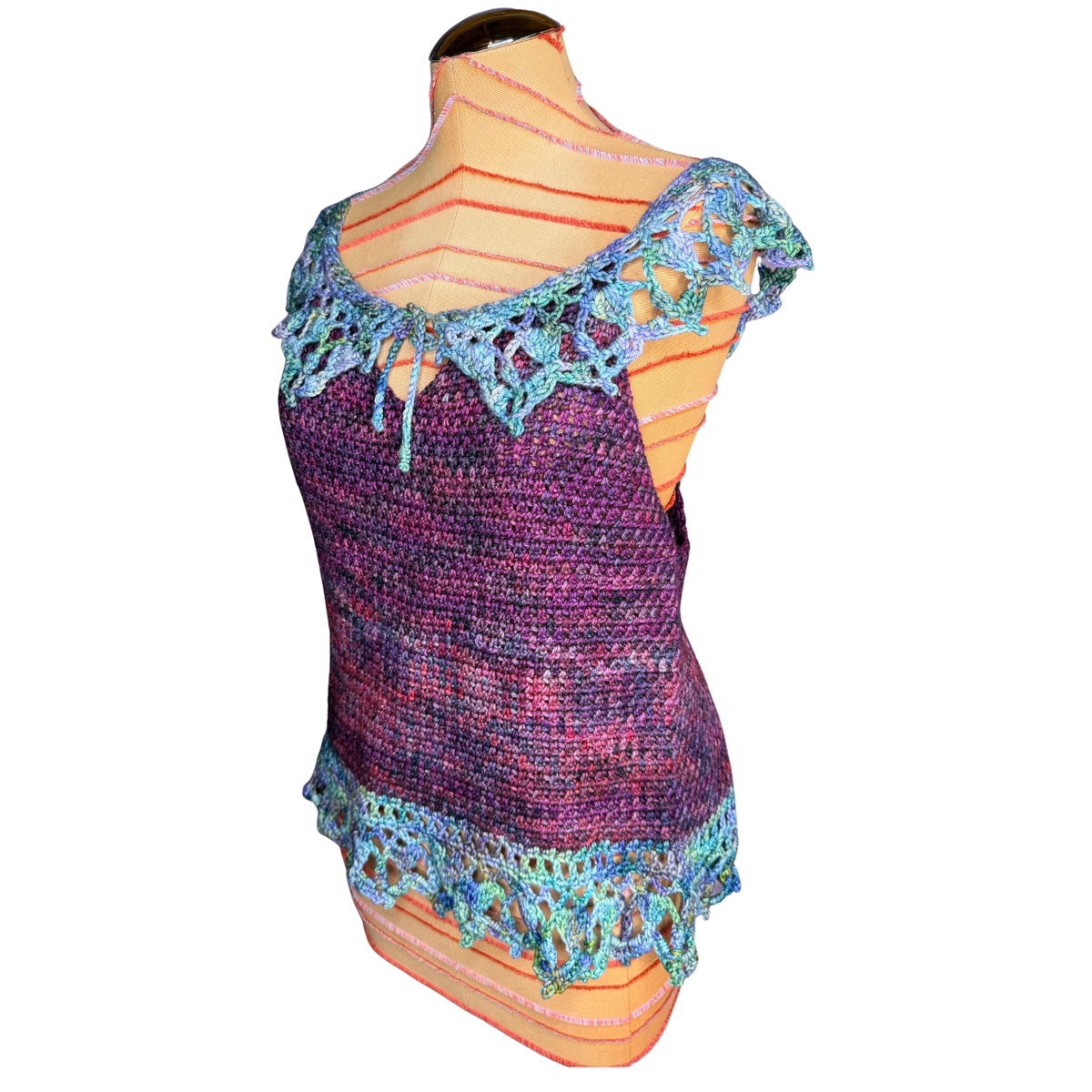 A hand-knit sleeveless top displayed on a striped mannequin torso against a white background. The top features a purple and pink marled body with blue and green lace trim at the shoulders and hem.