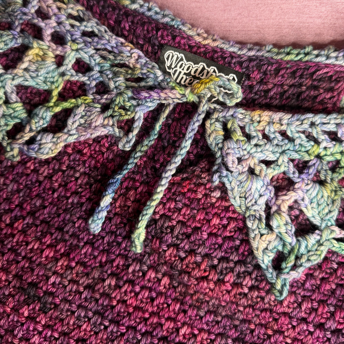 Close-up of a handmade crochet garment featuring a variegated multicolor yarn with shades of purple, pink, and green. The neckline is decorated with a lace trim tied with a bow, and a black fabric label with white script reading 'Woodshed theory' is stitched on the inside.