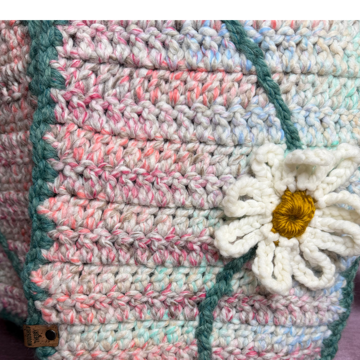 Close-up of a handmade crocheted textile with variegated pastel stripes in pink, blue, teal, and off-white, featuring a white daisy flower applique with a yellow center and a small woven brand tag near the bottom left.