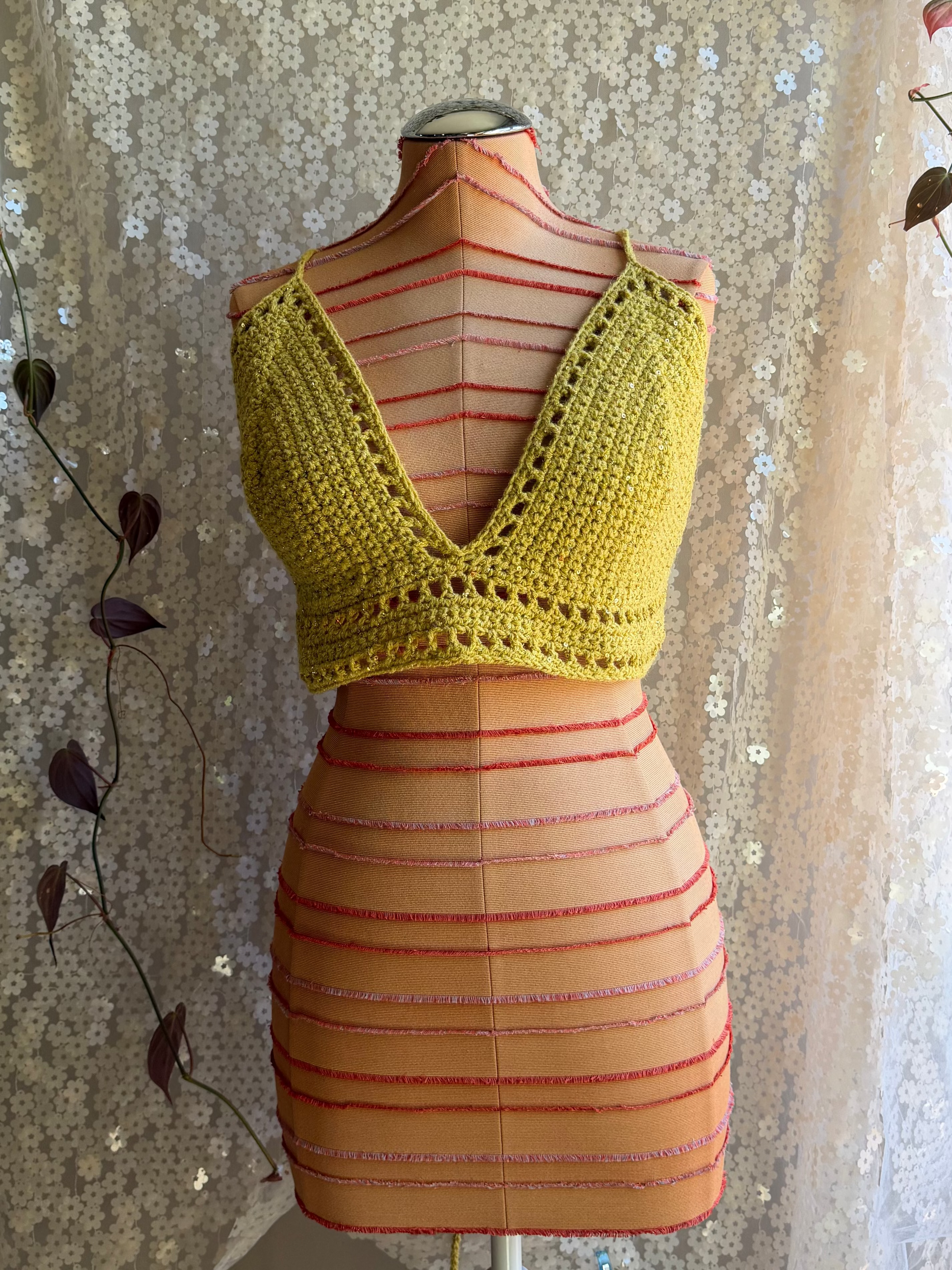 Mannequin wearing a yellow crochet top and striped skirt against a decorative white curtain.