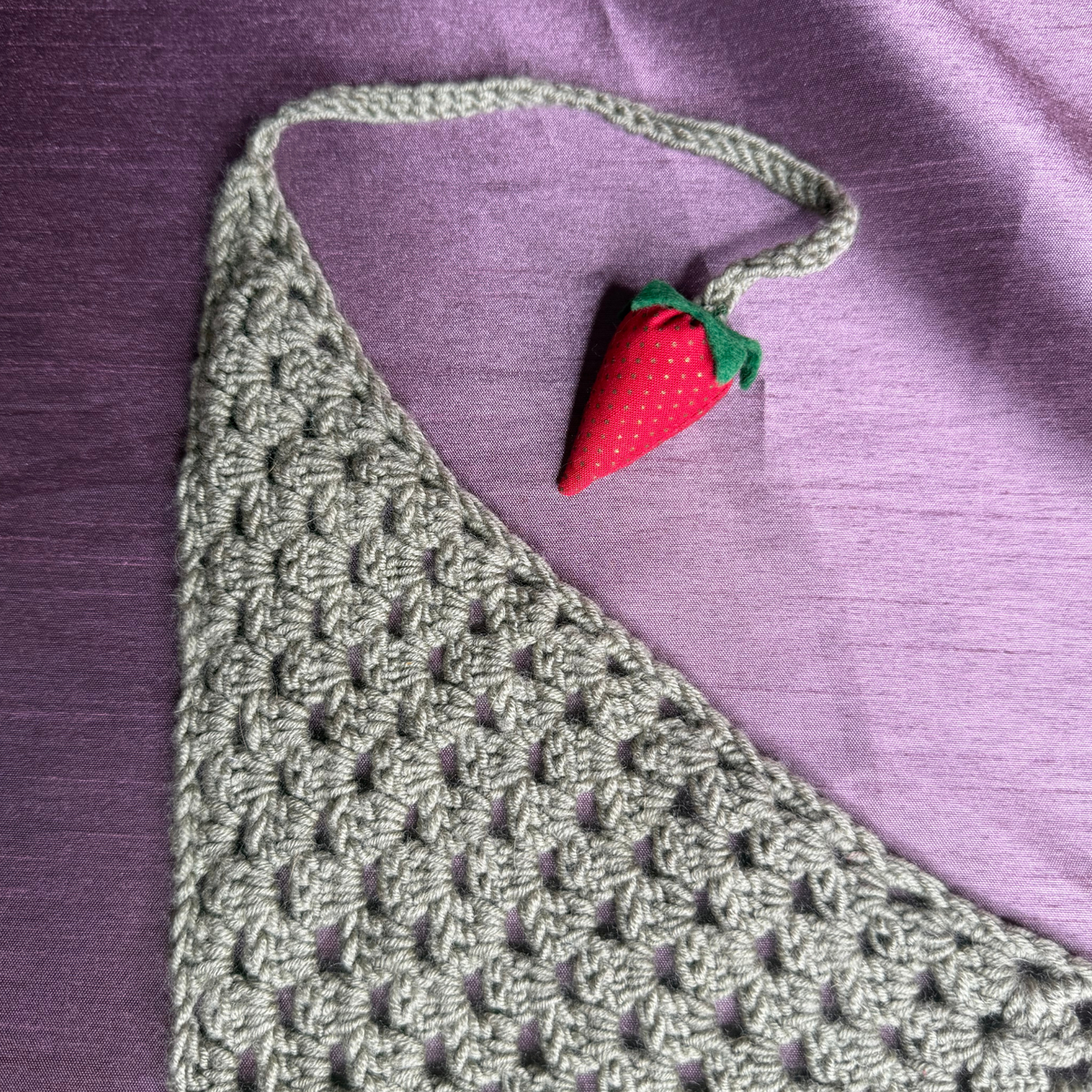 A close-up of a handmade crochet halter top in a sage green color, featuring a textured bobble stitch pattern and a small red strawberry charm with green leaves at the neck strap, set against a purple fabric background.