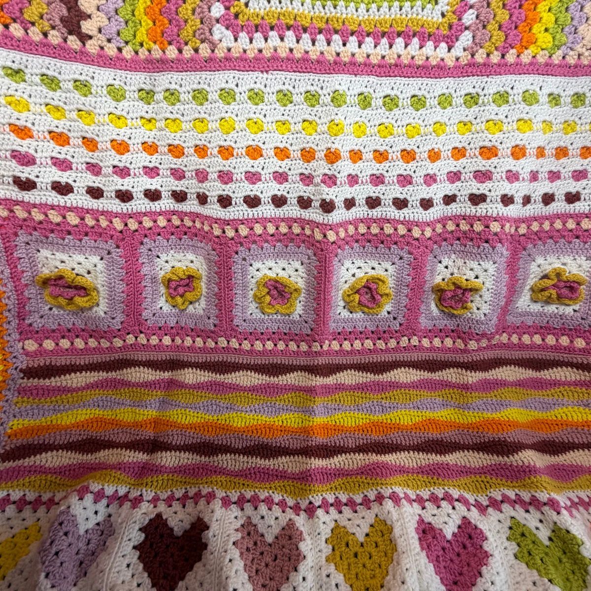 A close-up of a colorful crocheted blanket featuring striped bands, heart motifs, and floral granny square patterns in pink, yellow, orange, green, and white.