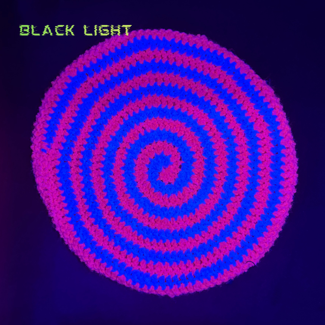 A circular spiral-patterned textile or mat shown under blacklight, glowing with alternating bright blue and pink-purple bands. The words 'BLACK LIGHT' appear in green text in the upper left corner.