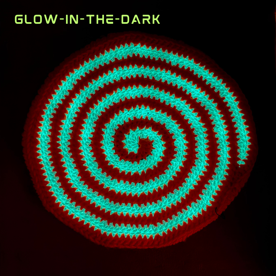 A circular spiral-shaped product, likely a mat or rug, shown glowing in the dark with bright cyan-green bands on a black background. The text 'GLOW-IN-THE-DARK' is displayed in the top-left corner.