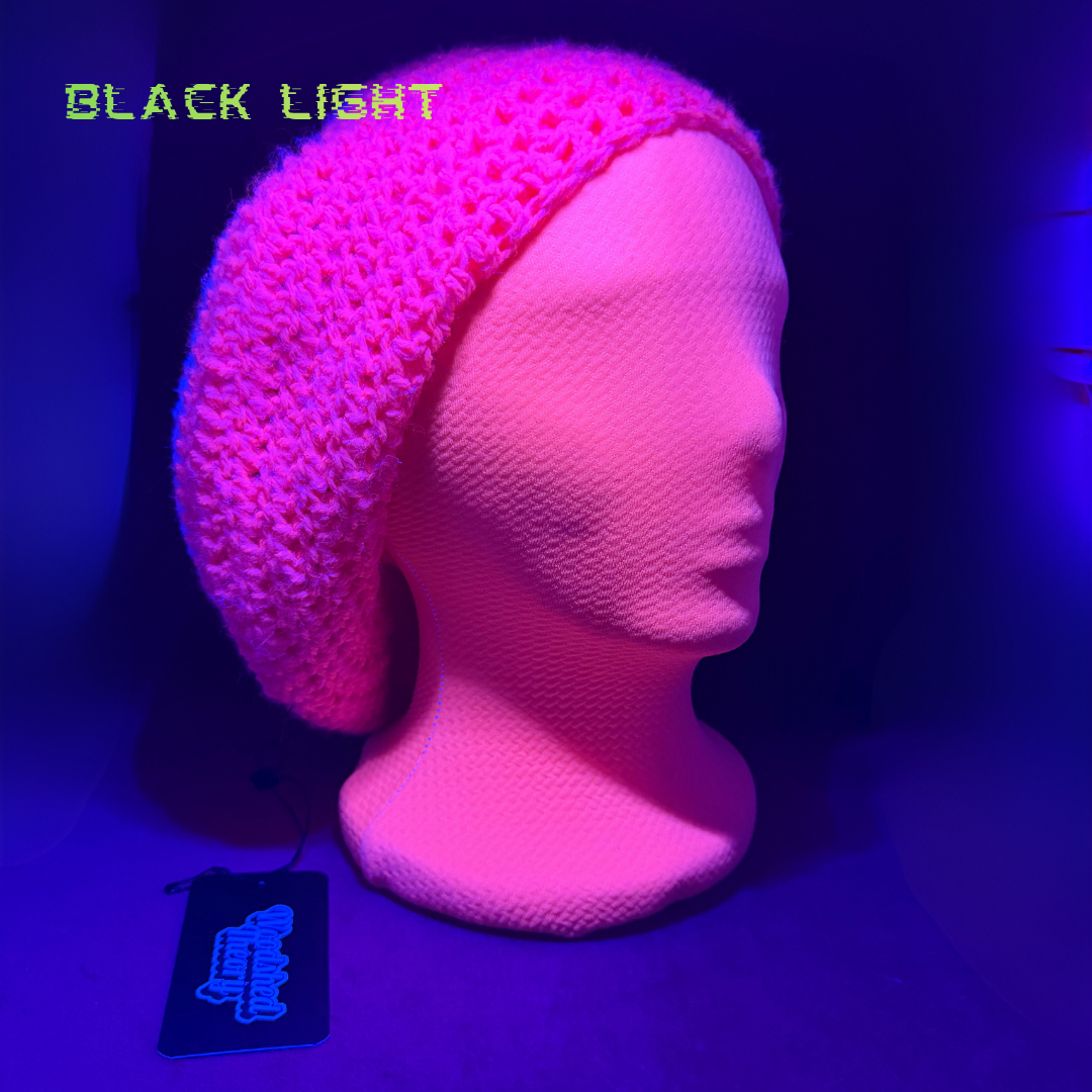 A pink crocheted beanie hat displayed on a mannequin head under blacklight, glowing bright pink under UV light. The words 'BLACK LIGHT' are written in green in the top left, and a black product tag is visible at the bottom left.