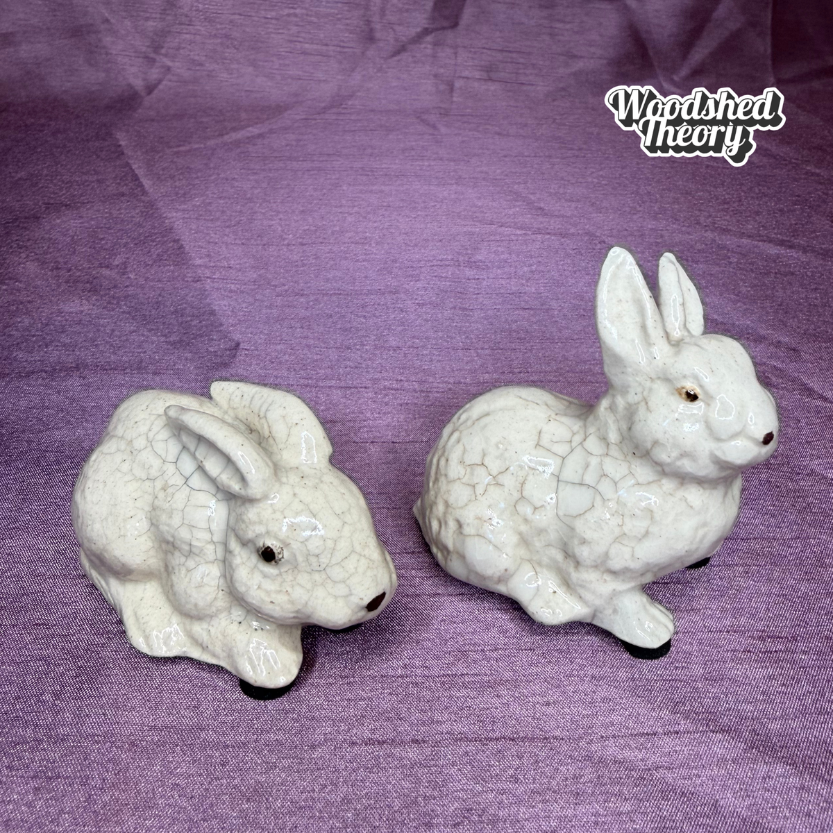 Two ceramic rabbit figurines with a glossy white glaze and crackle pattern, displayed on a textured purple fabric background. A 'Woodshed Theory' brand logo is visible in the top right corner.