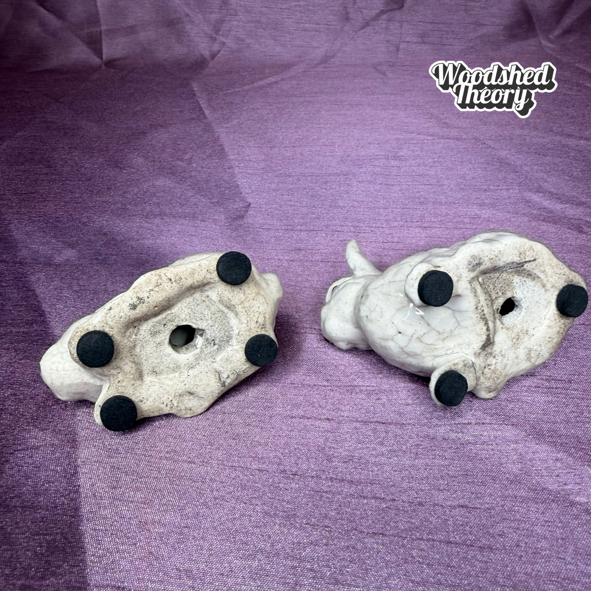 Two small white skull figurines with black circular feet, displayed on a textured purple fabric background. A 'Woodshed Theory' logo is visible in the top right corner.