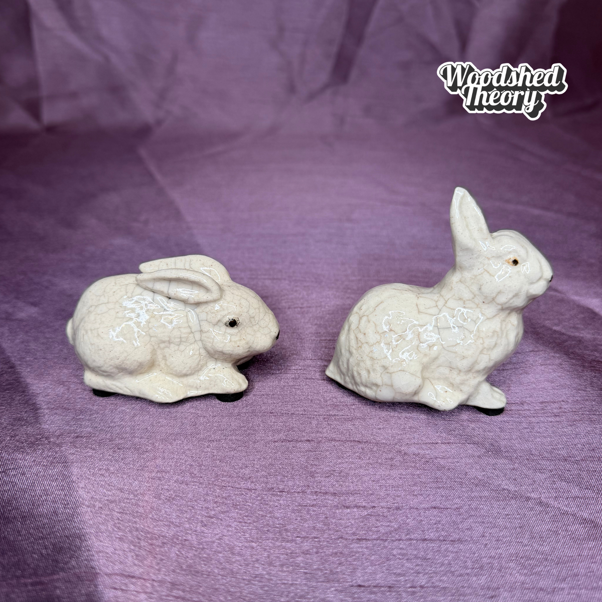 Two small ceramic rabbit figurines in an off-white glazed finish, displayed on a purple fabric backdrop. A 'Woodshed Theory' logo is visible in the top right corner.