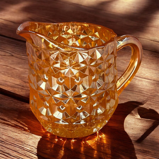 Vintage Jeannette Glass Company Buttons and Bows milk pitcher in iridescent amber, 1947–1949, with handle and shimmering finish. on table
